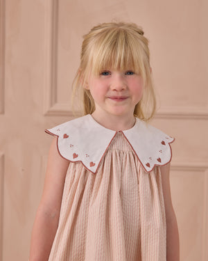 A young girl with blonde hair wears the Noralee Girls' Florence Dress, a vintage-inspired pink dress with a scalloped collar adorned with red hearts, standing against a beige background.