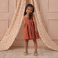 A young girl wears the rust-colored Noralee Girls' Betsy Dress with spaghetti straps, posing and smiling in front of beige curtains decorated with small red patterns.