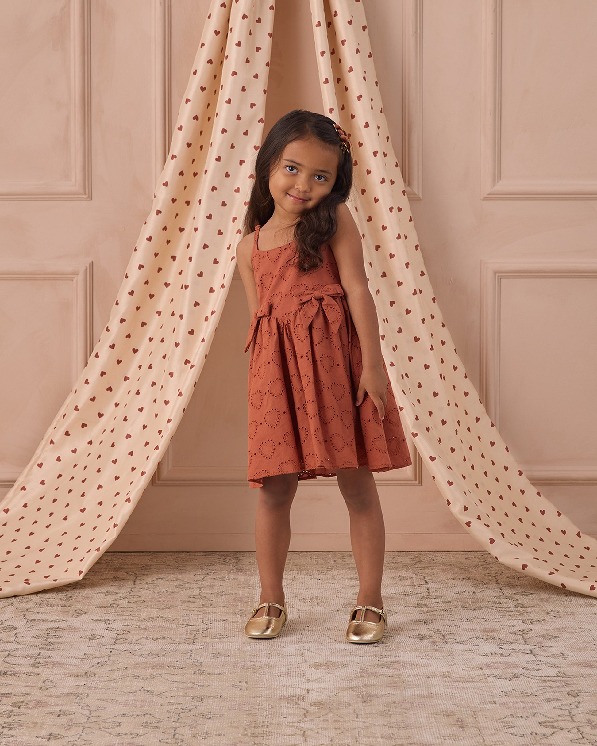 A young girl wears the rust-colored Noralee Girls' Betsy Dress with spaghetti straps, posing and smiling in front of beige curtains decorated with small red patterns.