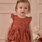 A toddler wears the Noralee Baby Girls' Betsy Romper—a rust-colored heart eyelet outfit with ruffle cap sleeves—while standing indoors by light-colored flowers, gazing slightly to the side.