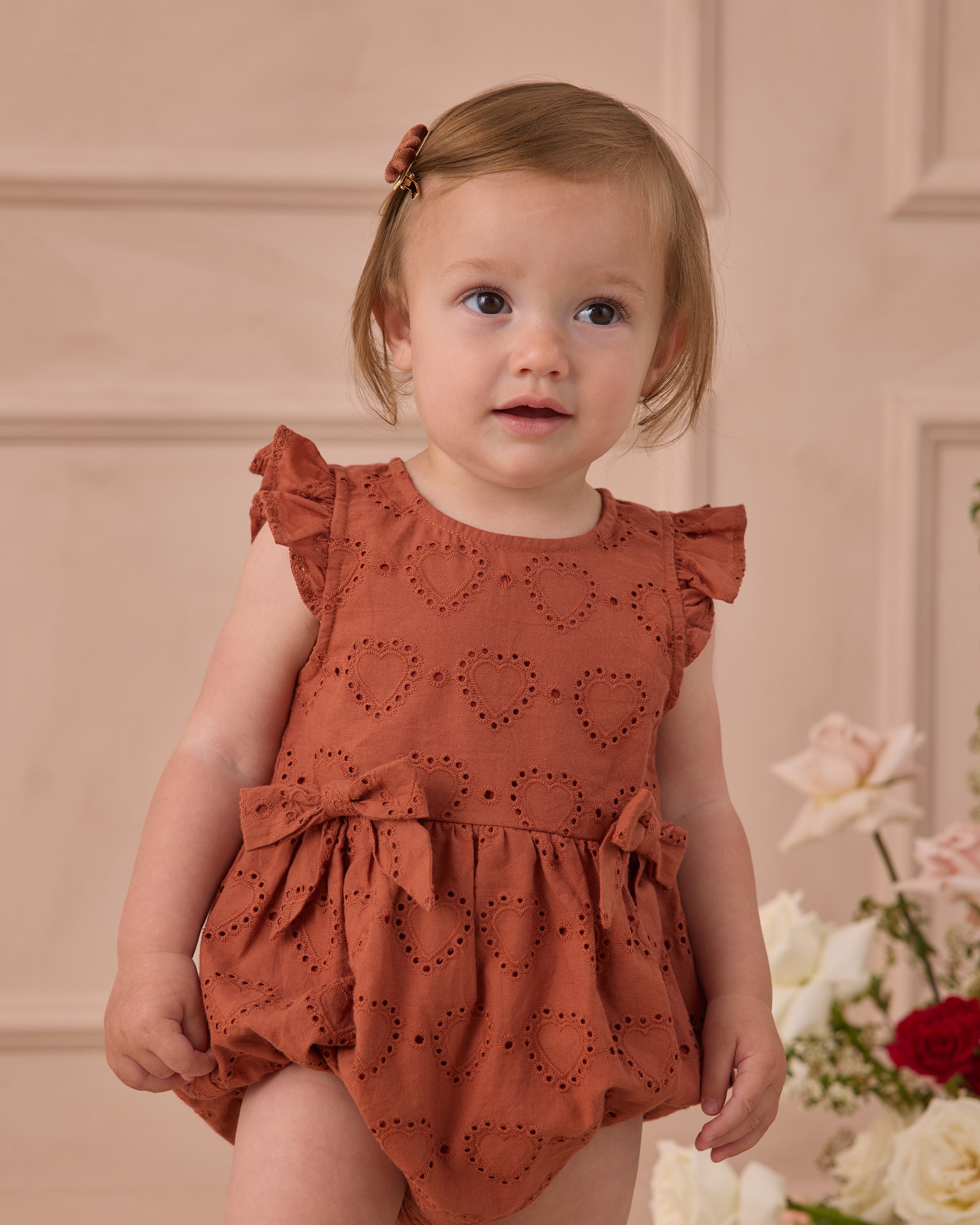 A toddler wears the Noralee Baby Girls' Betsy Romper—a rust-colored heart eyelet outfit with ruffle cap sleeves—while standing indoors by light-colored flowers, gazing slightly to the side.