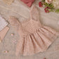The Noralee Baby Girls' & Girls' Valentina Tutu, a pink ruffled tulle dress with a smocked bodice and gold polka dots, is displayed on textured fabric with flowers, lace, and decorative stationery.
