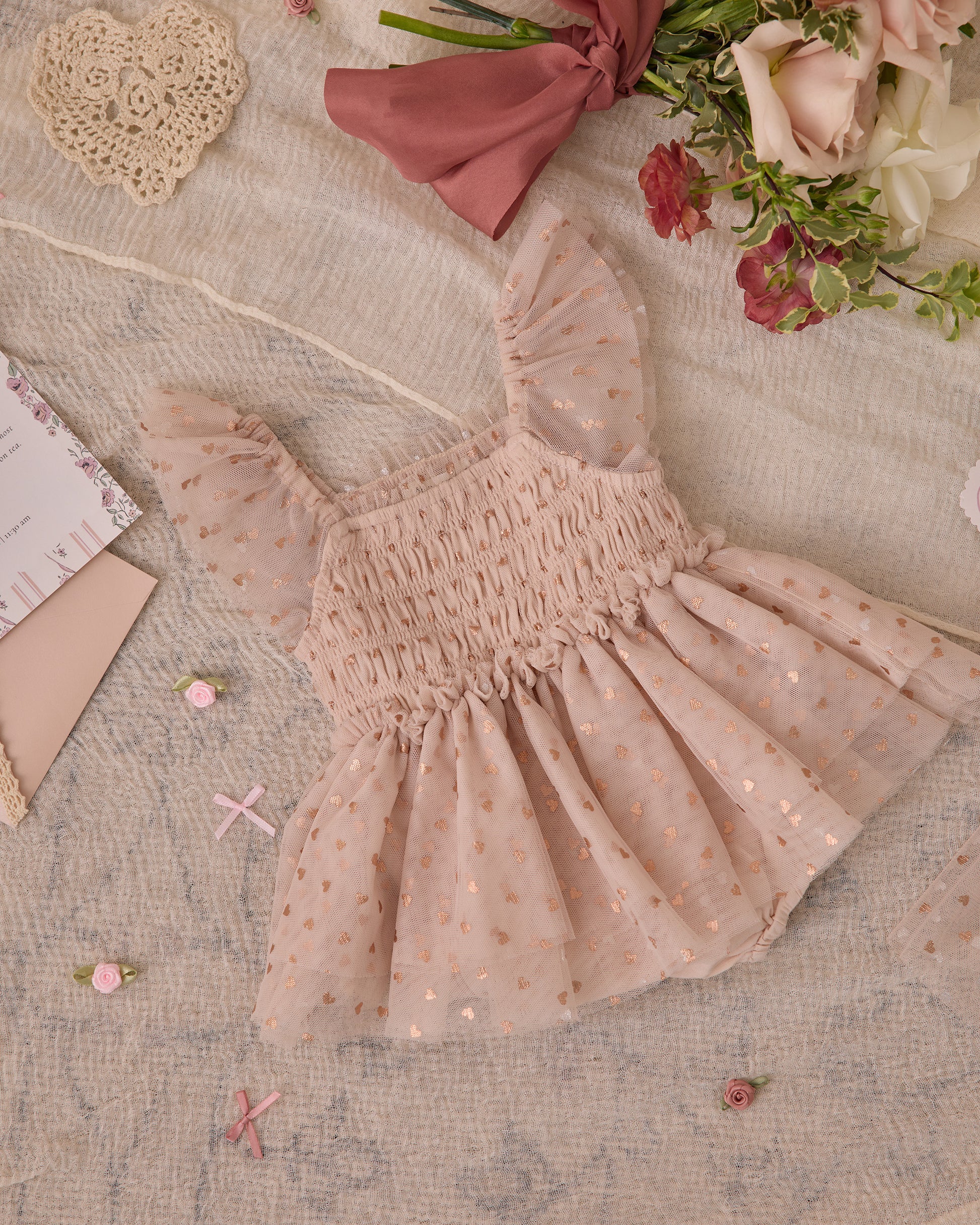 The Noralee Baby Girls' & Girls' Valentina Tutu, a pink ruffled tulle dress with a smocked bodice and gold polka dots, is displayed on textured fabric with flowers, lace, and decorative stationery.