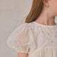 A child wearing the Noralee Girls’ Luna Dress—a cream-colored babydoll mini dress featuring puffed tulle sleeves and small metallic sequins—is shown from the shoulders up, turned to the side.