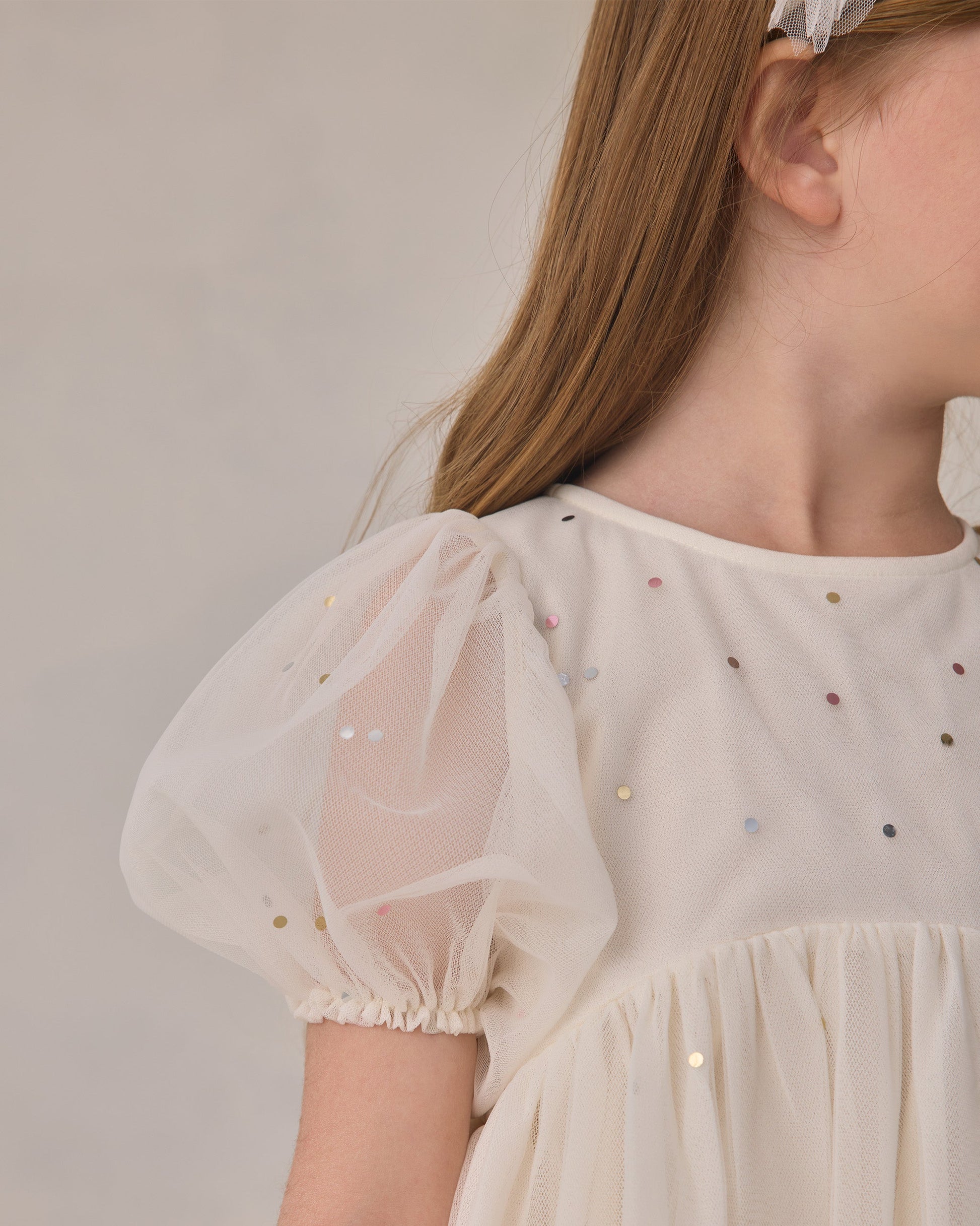 A child wearing the Noralee Girls’ Luna Dress—a cream-colored babydoll mini dress featuring puffed tulle sleeves and small metallic sequins—is shown from the shoulders up, turned to the side.