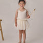 A young child wears the Noralee Baby Girls’ and Girls’ Lottie Tutu Set—a cream-colored fairy costume featuring a double-tiered tutu and flutter sleeve leotard with wings—while holding a star wand and standing beside a wooden stool against a neutral background.