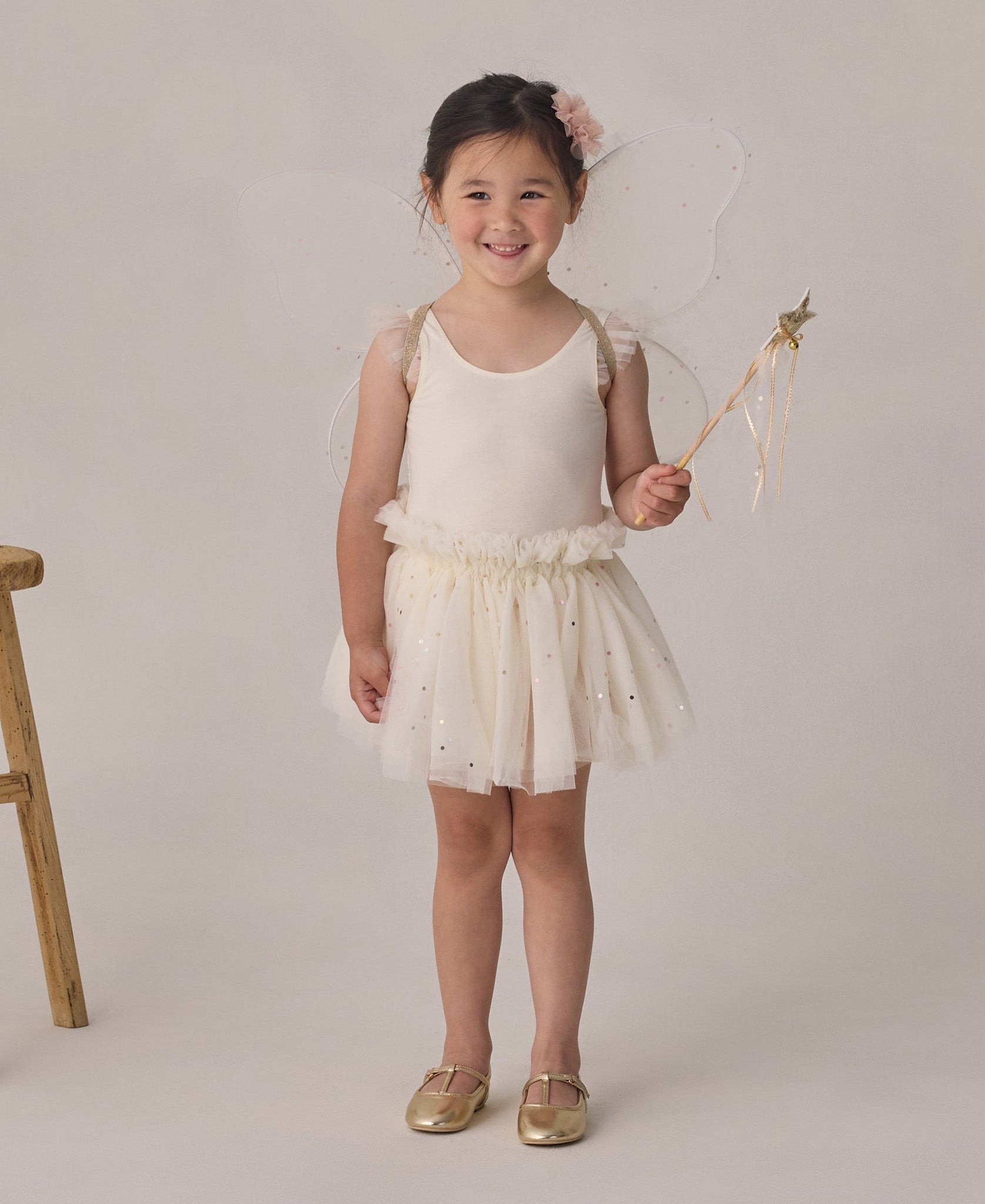 A young child wears the Noralee Baby Girls’ and Girls’ Lottie Tutu Set—a cream-colored fairy costume featuring a double-tiered tutu and flutter sleeve leotard with wings—while holding a star wand and standing beside a wooden stool against a neutral background.