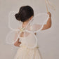 A child in a cream-colored dress with sparkles enjoys dress-up, wearing sheer butterfly wings and holding a wand from the Noralee Girls’ Fairy Set, facing away from the camera.