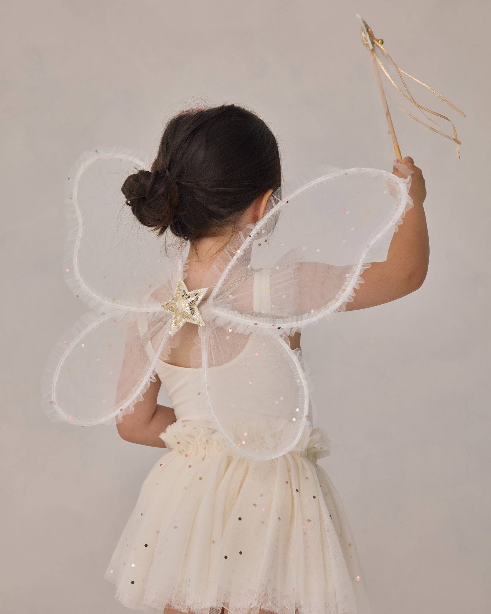 A child in a cream-colored dress with sparkles enjoys dress-up, wearing sheer butterfly wings and holding a wand from the Noralee Girls’ Fairy Set, facing away from the camera.