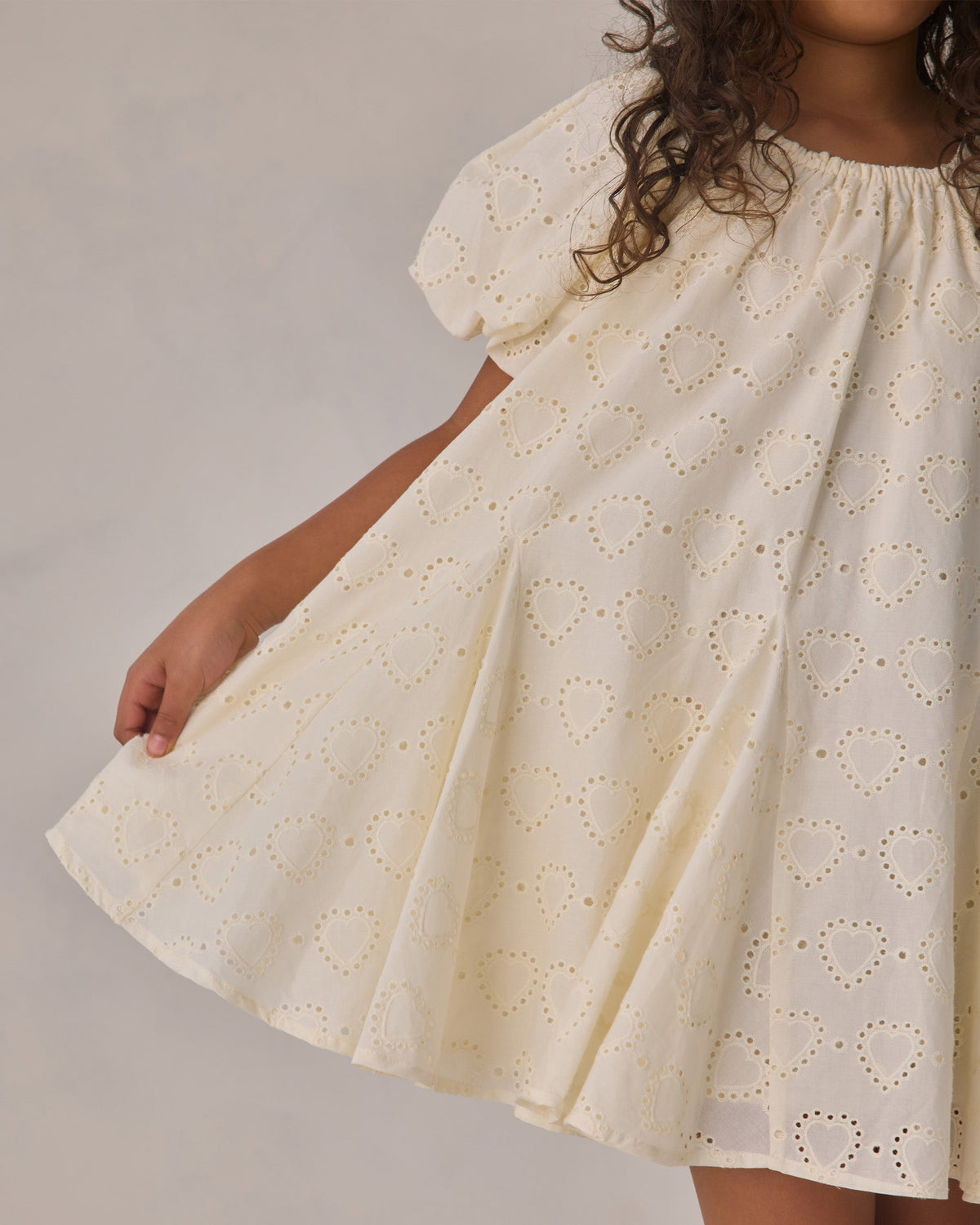 A child wearing the Noralee Baby Girls’ and Girls’ Maia Dress in off-white, featuring short sleeves and embroidered heart eyelet details, holding the skirt out to the side.