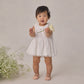 Wearing the Noralee Girls’ Georgia Dress—a charming white mini dress with delicate eyelet detailing—a toddler holds a white flower and stands beside a cluster of small white blooms against a simple backdrop, creating an ideal look for any special occasion.