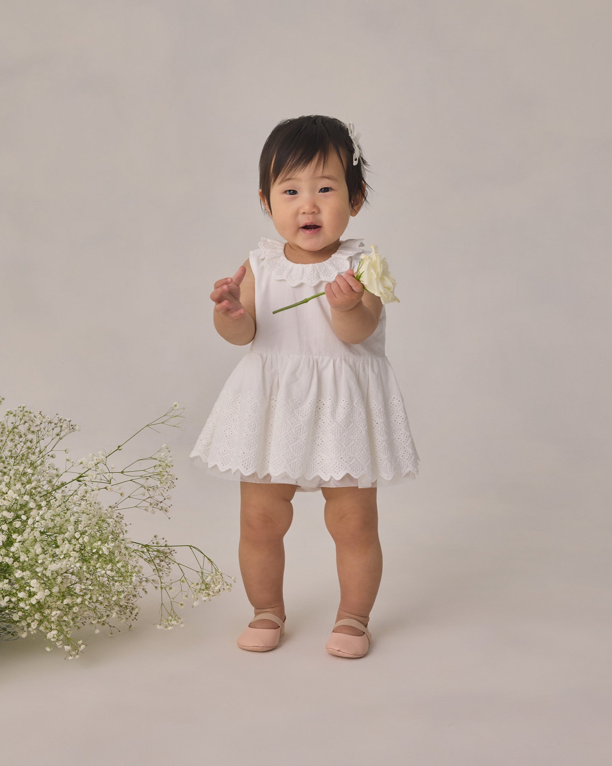 Wearing the Noralee Girls’ Georgia Dress—a charming white mini dress with delicate eyelet detailing—a toddler holds a white flower and stands beside a cluster of small white blooms against a simple backdrop, creating an ideal look for any special occasion.