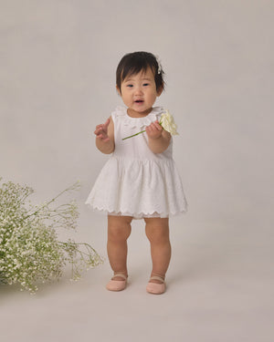 Wearing the Noralee Girls’ Georgia Dress—a charming white mini dress with delicate eyelet detailing—a toddler holds a white flower and stands beside a cluster of small white blooms against a simple backdrop, creating an ideal look for any special occasion.