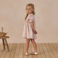 A young girl in a vintage mini dress with delicate floral embroidery and a smocked neckline stands on a wooden floor, looking over her shoulder. A small bench with a basket is on her left.