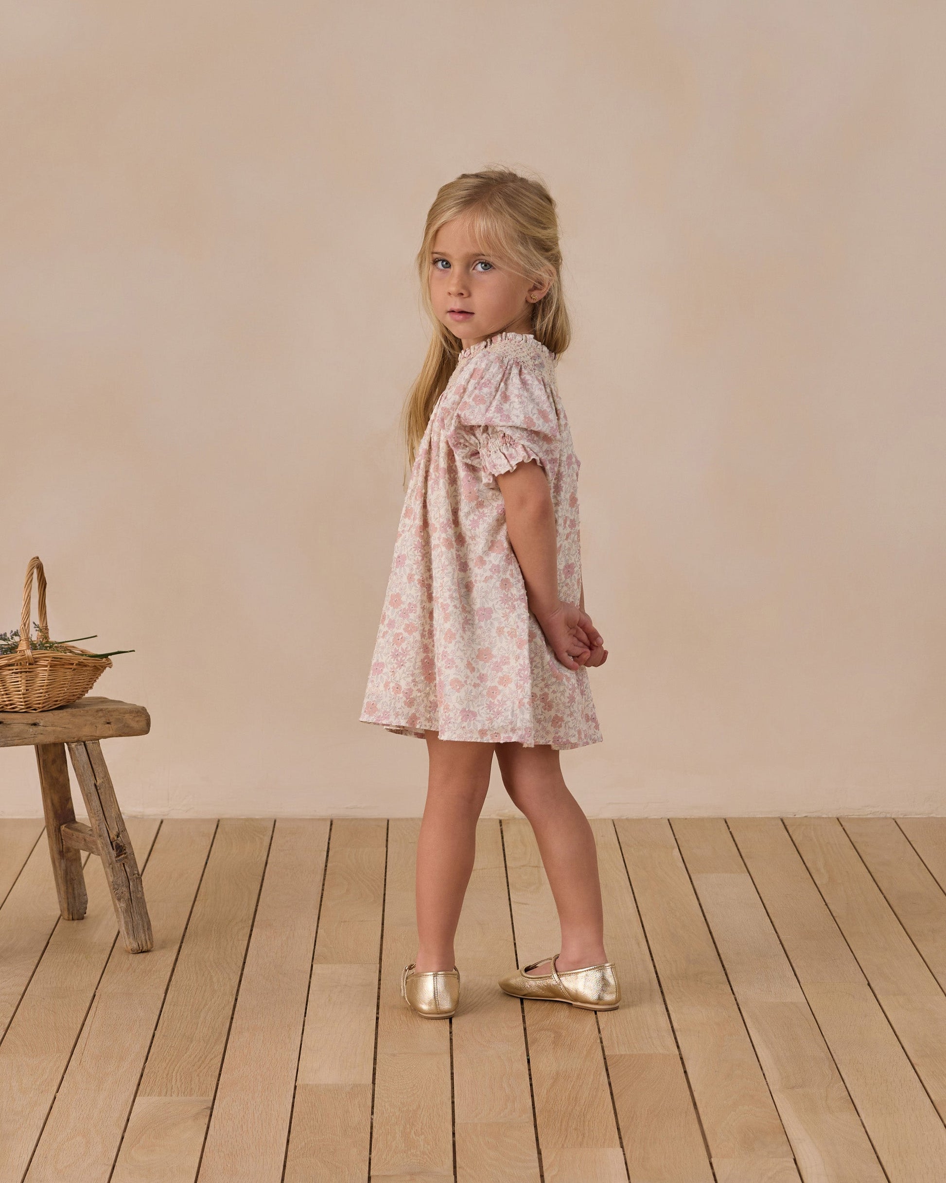 A young girl in a vintage mini dress with delicate floral embroidery and a smocked neckline stands on a wooden floor, looking over her shoulder. A small bench with a basket is on her left.