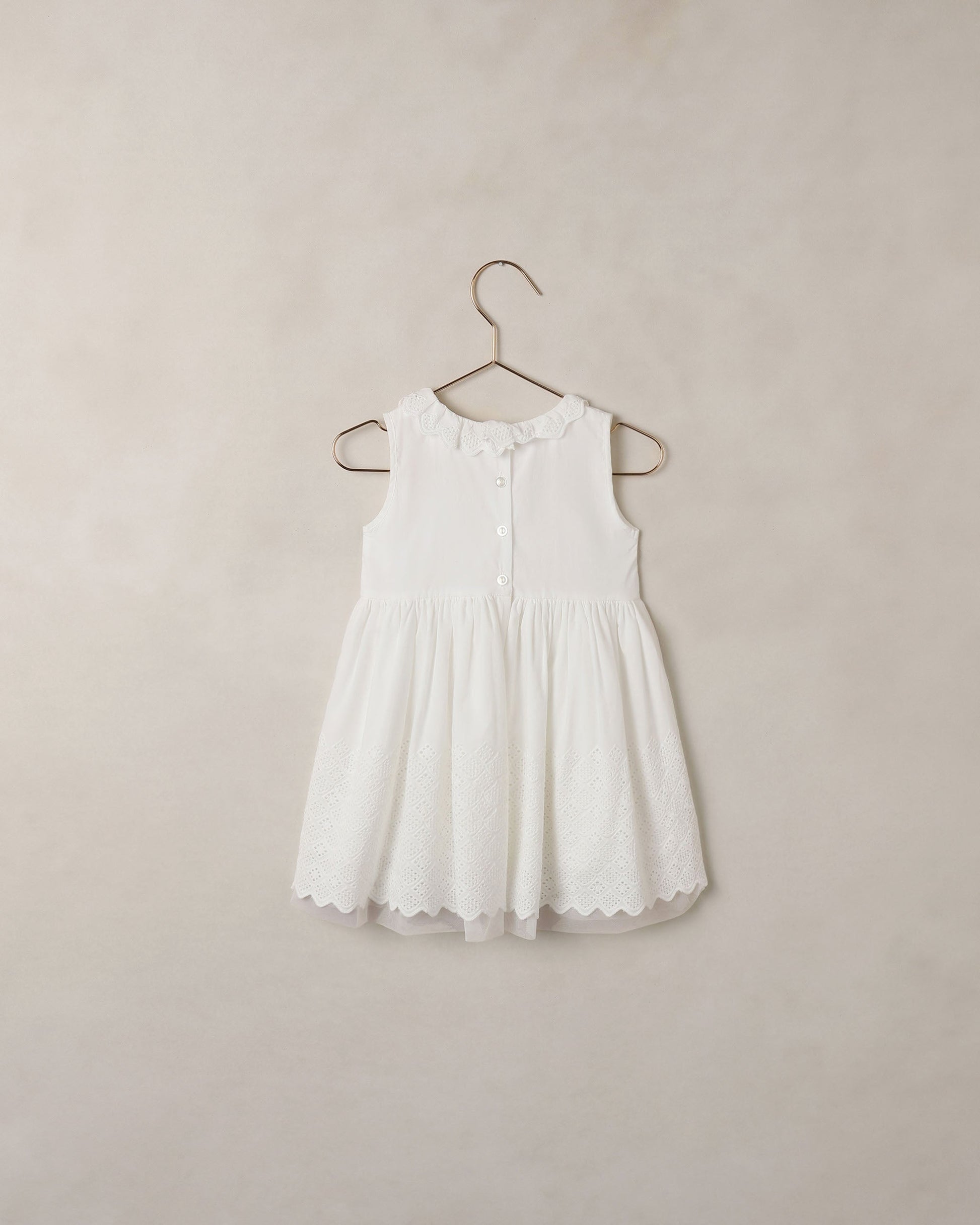 The Noralee Girls’ Georgia Dress is a sleeveless white baby mini dress featuring decorative buttons, delicate eyelet detailing, and lace trim, shown hanging on a metal hanger against a simple light background—perfect for special occasions.