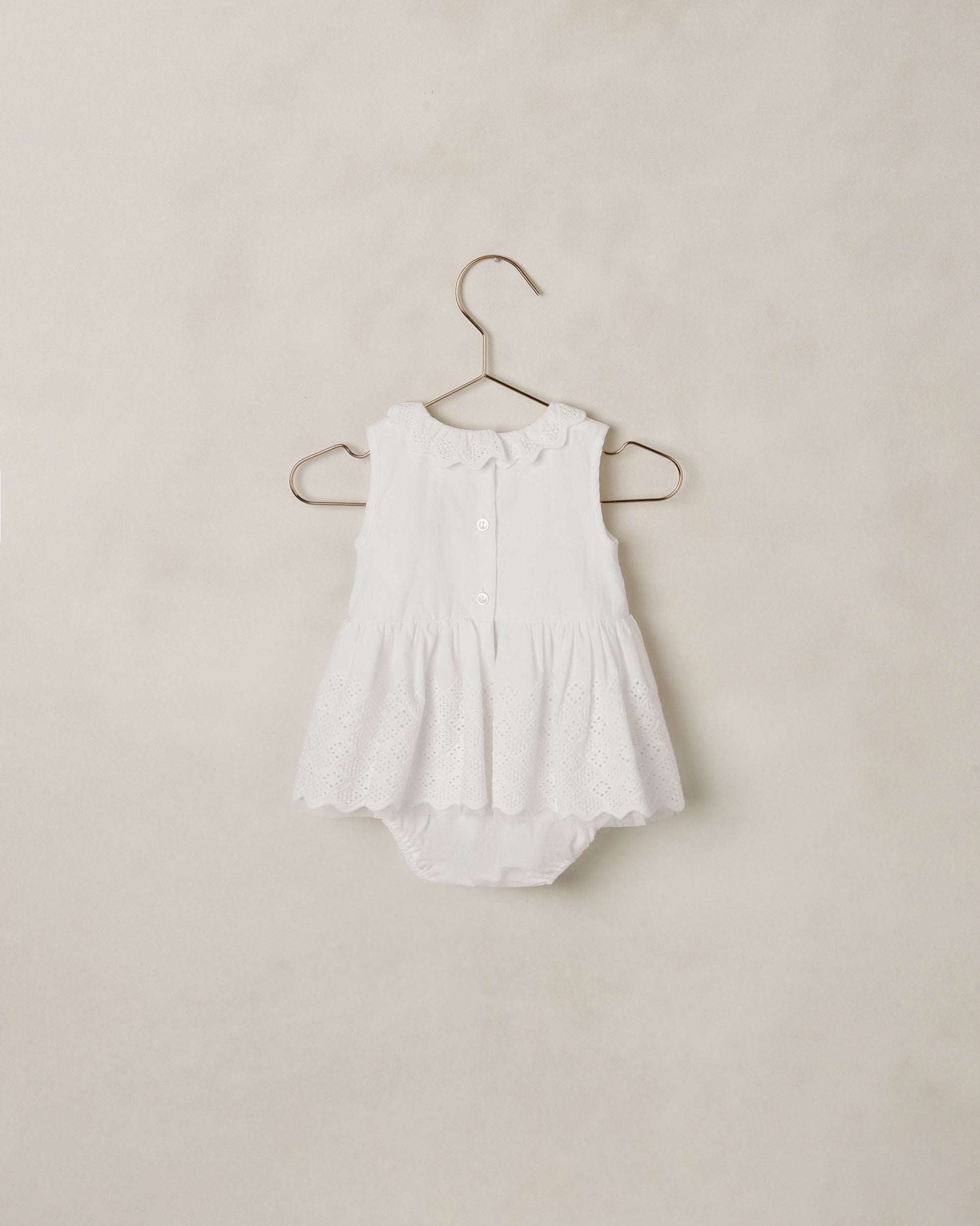 The Noralee Baby Girls’ Georgia Romper features a white sleeveless design with a scalloped neckline, ruffled collar, eyelet lace skirt detail, and button front for easy changing, displayed on a hanger against a neutral background.