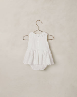The Noralee Baby Girls’ Georgia Romper features a white sleeveless design with a scalloped neckline, ruffled collar, eyelet lace skirt detail, and button front for easy changing, displayed on a hanger against a neutral background.