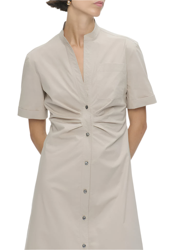 A person models the Callas Milano Noor Dress, highlighting its flattering fit. The beige, short-sleeved, button-up shirt dress features a mandarin collar and an elegant midi-length design, adding sophistication with hands placed behind their back.