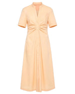 The Noor Dress by Callas Milano is a peach fit and flare midi shirt dress featuring short sleeves, a v-neck, button-down front, and ruched waist detail for a flattering silhouette.