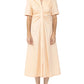 A woman stands against a plain white background wearing the Noor Dress by Callas Milano, a light peach fit and flare midi shirt dress with short sleeves and gathered waist detail, paired with black sandals.
