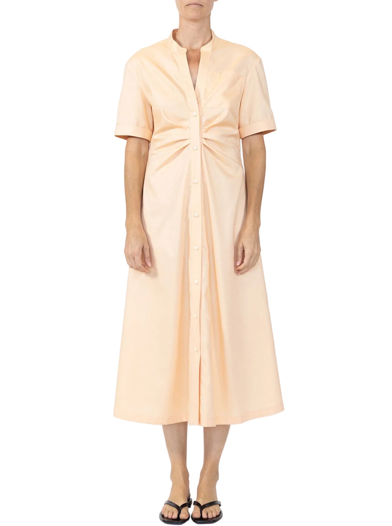A woman stands against a plain white background wearing the Noor Dress by Callas Milano, a light peach fit and flare midi shirt dress with short sleeves and gathered waist detail, paired with black sandals.