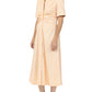 A person wears the Noor Dress by Callas Milano—a light peach, button-front midi shirt dress with short sleeves and a gathered front—paired with black flip-flops, standing against a plain background.