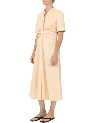 A person wears the Noor Dress by Callas Milano—a light peach, button-front midi shirt dress with short sleeves and a gathered front—paired with black flip-flops, standing against a plain background.
