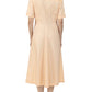 A person stands facing away, wearing the Noor Dress by Callas Milano—a light peach, short-sleeve, midi shirt dress with a fitted waist and flared skirt.
