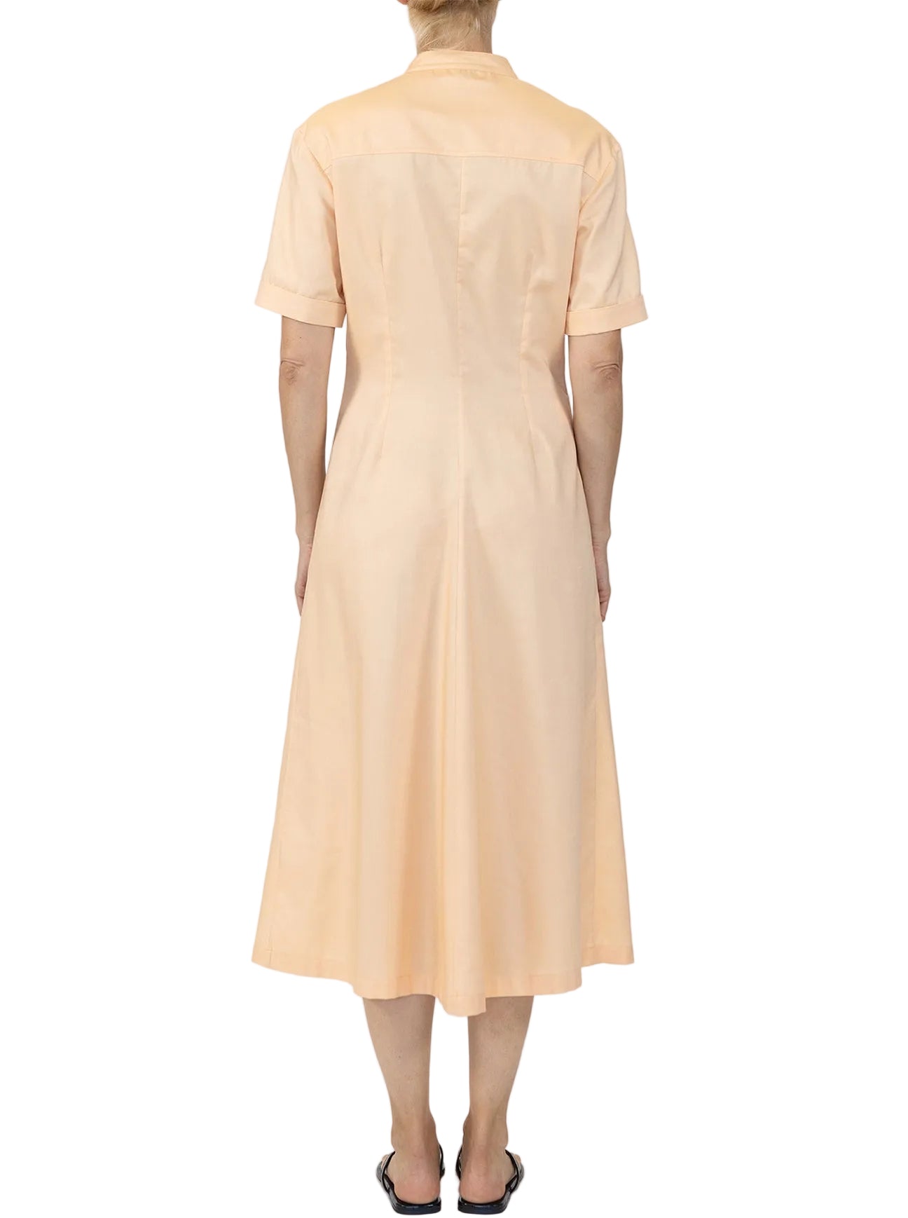 A person stands facing away, wearing the Noor Dress by Callas Milano—a light peach, short-sleeve, midi shirt dress with a fitted waist and flared skirt.