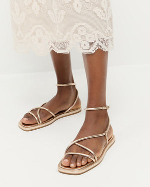 A person wearing Loeffler Randall Noor Strappy Ankle Wrap Flat sandals in champagne metallic and a white lace midi skirt, standing on a white floor.