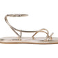 The Noor Strappy Ankle Wrap Flat by Loeffler Randall is a champagne metallic sandal crafted from Italian leather, featuring thin straps, an ankle wrap, toe loop, and showcased on a white background.