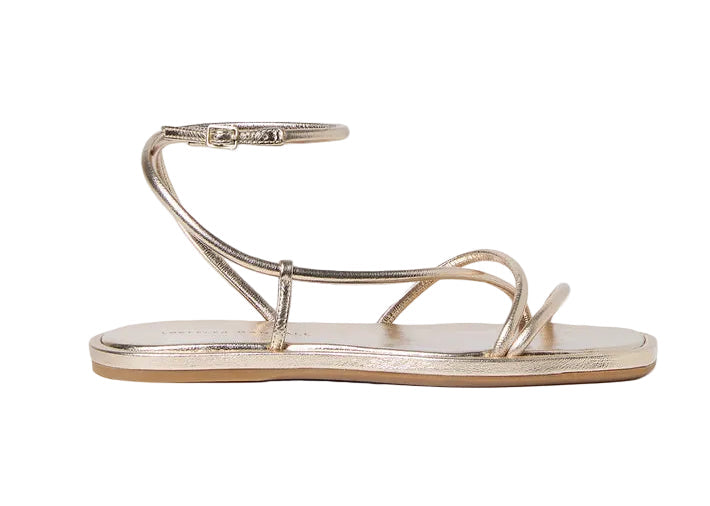 The Noor Strappy Ankle Wrap Flat by Loeffler Randall is a champagne metallic sandal crafted from Italian leather, featuring thin straps, an ankle wrap, toe loop, and showcased on a white background.