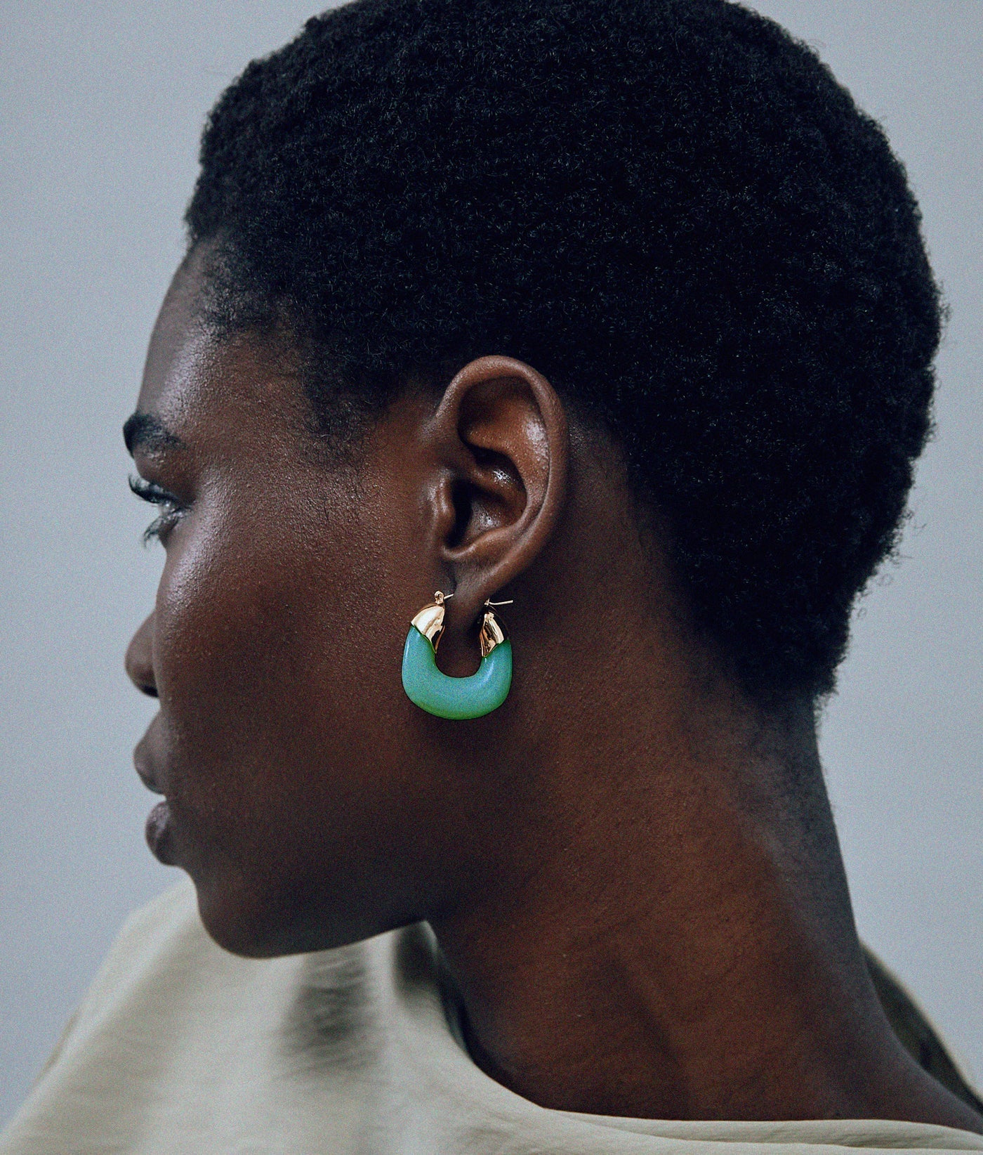 A person with short, curly hair is shown in profile wearing Lizzie Fortunato Organic Mini Gold Hoops. The background is plain and neutral.