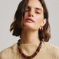 A woman with shoulder-length brown hair wears a cream textured top, chunky amber and brown beaded necklace, and Lizzie Fortunato Organic Gold Hoops, looking directly at the camera.