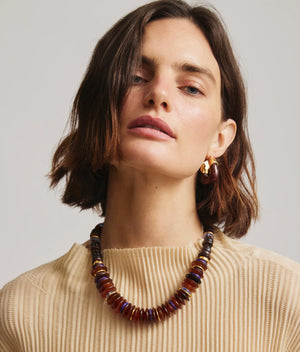 A woman with shoulder-length brown hair wears a cream textured top, chunky amber and brown beaded necklace, and Lizzie Fortunato Organic Gold Hoops, looking directly at the camera.