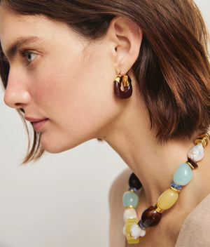 A woman with short brown hair wears a chunky beaded necklace in blue, yellow, and brown by Lizzie Fortunato, paired with Lizzie Fortunato's Organic Mini Gold Hoops.