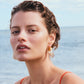A woman with wet, slicked-back hair wears Lizzie Fortunato’s Organic Gold Hoops—large gold and white resin earrings—and a thin-strap orange top as she stands by the ocean.