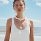 A woman in a white swimsuit, shell jewelry, and Lizzie Fortunato Organic Mini Gold Hoops stands on a beach with the ocean and blue sky behind her.