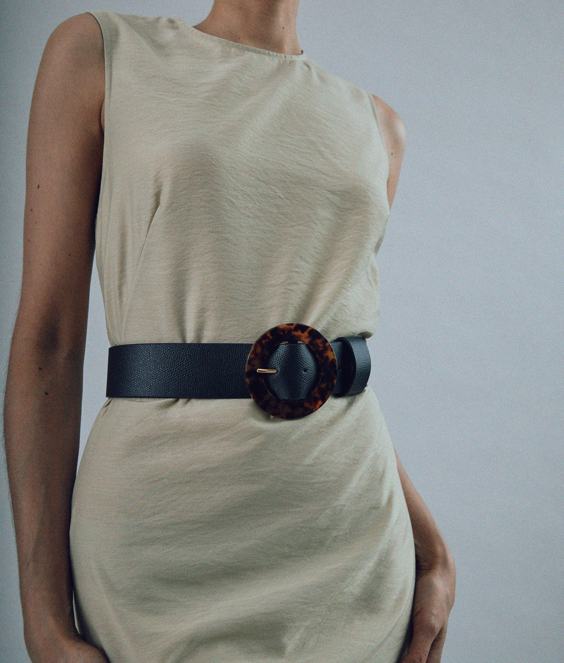 A person in a sleeveless beige dress wears the Lizzie Fortunato Louise Tortoise Acrylic Buckle Belt featuring an oversized tortoiseshell buckle.