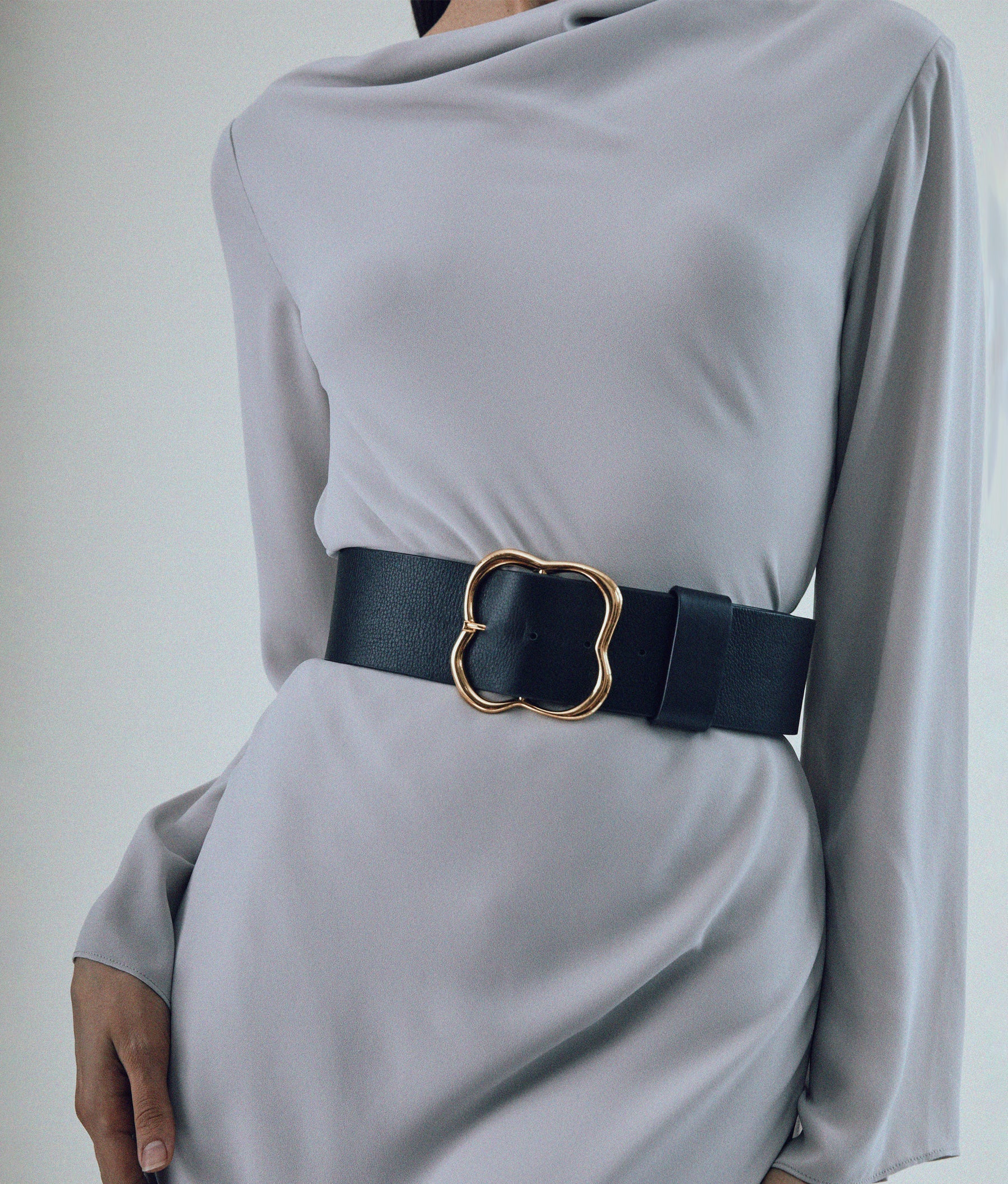 A person in a light gray dress cinched with the wide Lizzie Fortunato Florence Gold Buckle Belt featuring an ornate gold-plated buckle.