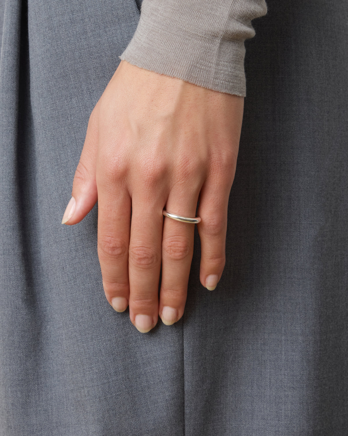 A hand with short, natural nails wears the Nanna Ring by Lie Studio, a classic curved sterling silver band, on the ring finger against a gray fabric background.