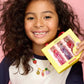 A smiling, curly-haired child wears a colorful beaded necklace and holds a yellow box labeled "Super Smalls" with the Gloss it Fruity Lip Gloss Ring Set, against a pink background.