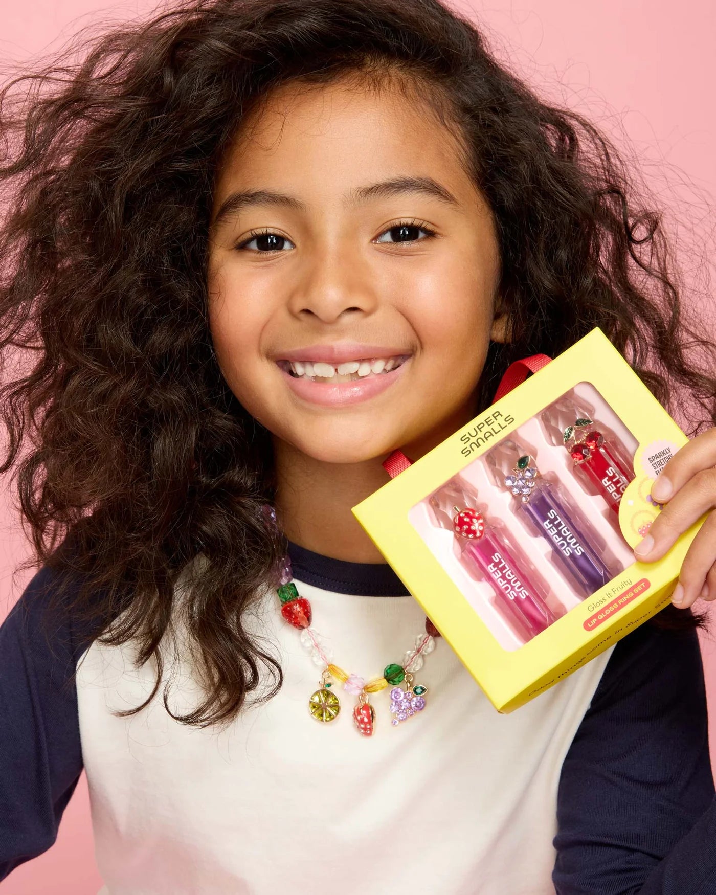 A smiling, curly-haired child wears a colorful beaded necklace and holds a yellow box labeled "Super Smalls" with the Gloss it Fruity Lip Gloss Ring Set, against a pink background.