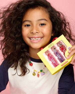 A smiling, curly-haired child wears a colorful beaded necklace and holds a yellow box labeled "Super Smalls" with the Gloss it Fruity Lip Gloss Ring Set, against a pink background.