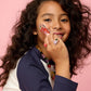 A young girl with long, curly hair wears a navy and white shirt and colorful jewelry, smiling brightly against a pink background as she holds a red lipstick to her cheek. She accessorizes with the Super Smalls Gloss it Rainbow Lip Gloss Ring Set, adding an extra pop of fun to her radiant look.