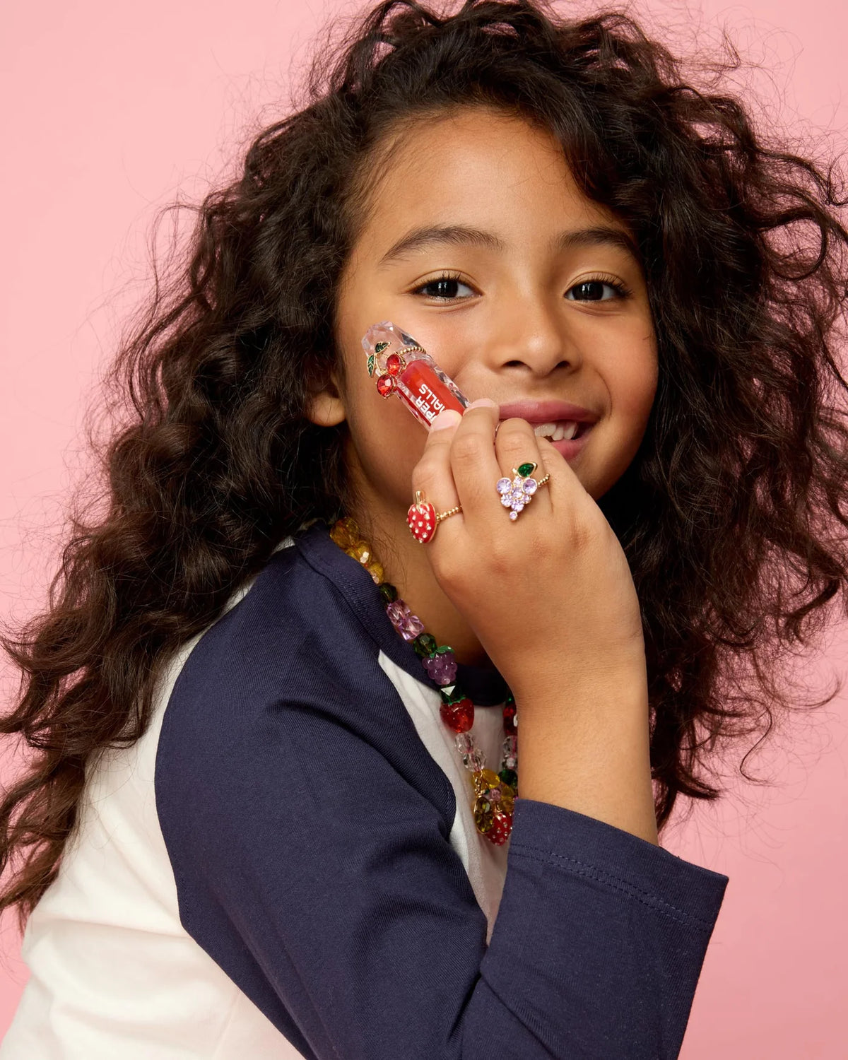 A young girl with long, curly hair wears a navy and white shirt and colorful jewelry, smiling brightly against a pink background as she holds a red lipstick to her cheek. She accessorizes with the Super Smalls Gloss it Rainbow Lip Gloss Ring Set, adding an extra pop of fun to her radiant look.