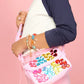 A person wearing a white and navy shirt, pink pants, and colorful bracelets holds a vibrant DIY gem purse—created with the Super Smalls Preppy Purse Gemify™ Craft Kit—adorned with large, multicolored plastic gems against a pink background.