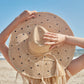 With long blonde hair, a person faces the beach under a clear blue sky, holding the Mar Y Sol Natalia Hat—a polka dot sun hat with a wide brim.