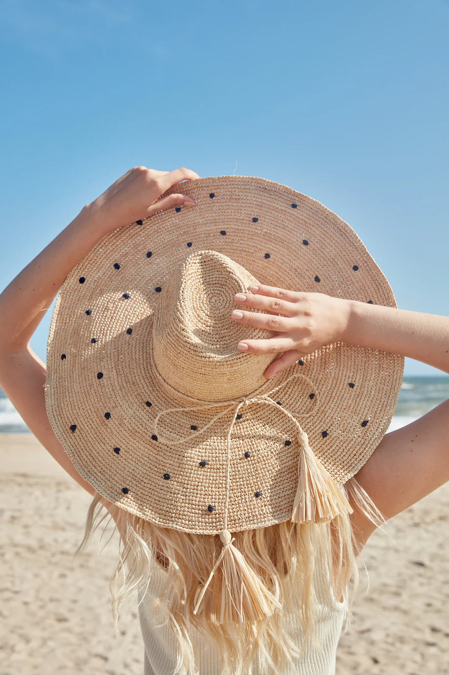 With long blonde hair, a person faces the beach under a clear blue sky, holding the Mar Y Sol Natalia Hat—a polka dot sun hat with a wide brim.